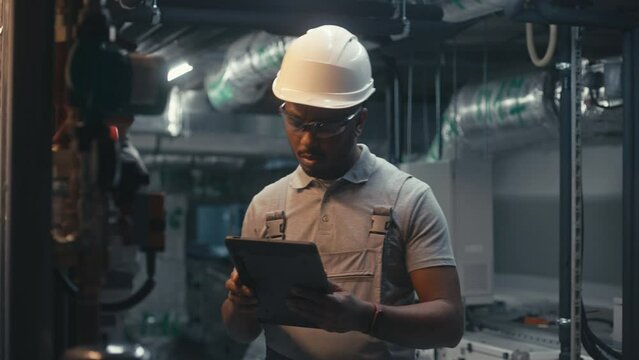 Male technician worker wearing safety uniform and hard hat works using tablet computer. African American inspector checks pipeline system on modern factory, plant or industrial energy facility.