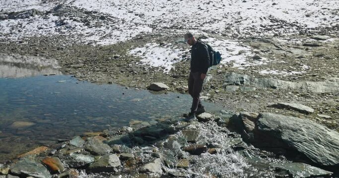Male hiker crosses mountain stream by using stepstones, reflection