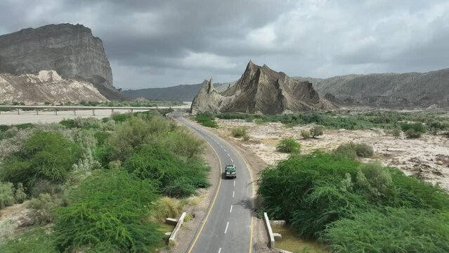 Aerial drone following shot over a car driving along the highway surrounded by mountains in Hingol, Balochistan, Pakistan on a cloudy day.