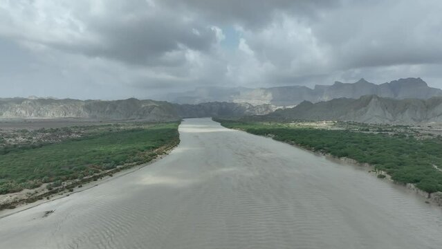 Aerial drone tilt up shot over dried riverbed surrounded by green vegetation with mountain range in the background in Hingol, Balochistan, Pakistan at daytime.