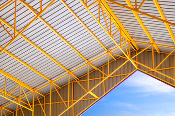 Fototapeta premium Low angle view of yellow metal roof beams and aluminum corrugated tile roof of industrial building with blue sky, inside view with copy space