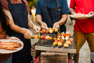Skewers with colorful vegetables and meat grilling, family and friends gathering in the background, enjoying a sunny outdoor meal. BBQ party, barbeque grill weekend, happy family.