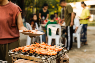 Skewers with colorful vegetables and meat grilling, family and friends gathering in the background, enjoying a sunny outdoor meal. BBQ party, barbeque grill weekend, happy family.