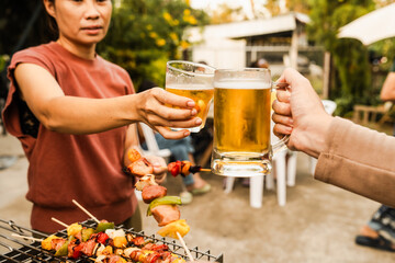 Outdoor barbecue with various meats on grill, two people holding plates and drinks, smoke rising,...