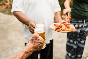 Outdoor barbecue with various meats on grill, two people holding plates and drinks, smoke rising, festive and casual atmosphere. holding glass of beer. family party.