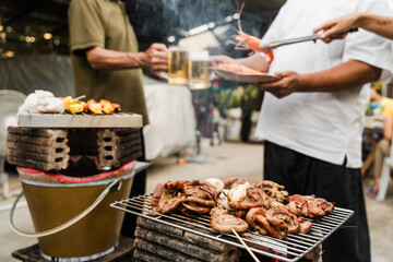 Outdoor barbecue with various meats on grill, two people holding plates and drinks, smoke rising, festive and casual atmosphere. holding glass of beer. family party.