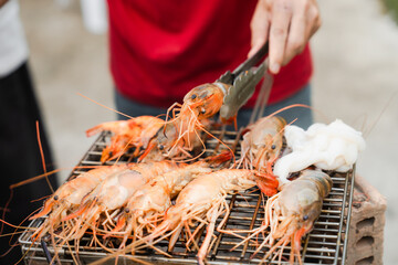 Grilling shrimp on a barbecue. Someone in a red shirt is using tongs to flip the shrimp, with smoke rising.