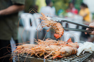 Grilling shrimp on a barbecue. Someone in a red shirt is using tongs to flip the shrimp, with smoke rising.