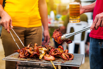 Barbecue with grilled chicken and sausages. People in casual clothes are cooking and holding a glass of beer.