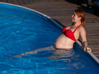 A red-haired pregnant woman in a red bikini is resting in the pool, leaning on the side.