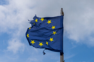 A torn EU flag blows in the wind with a few clouds in the background