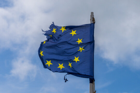 A torn EU flag blows in the wind with a few clouds in the background