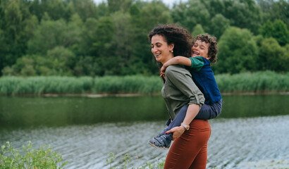 Fototapeta na wymiar Happy little curly boy piggyback on mom against river and forest on background. Cheerful hispanic woman entertains son on nature at holiday vacations. Childhood, family time.
