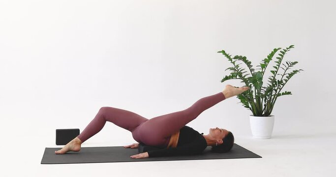 Athletic woman performs leg rolls with one brick under her lower back, an exercise for mastering the longitudinal splits, training in leggings and a long sleeve top on a mat in the studio