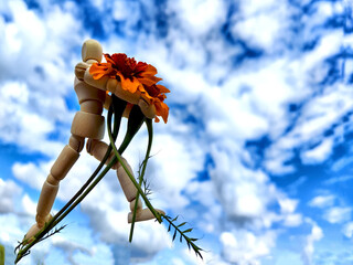 Wooden toy man with flowers and sky with white clouds on backgroudn. Concept of holiday, bouquet,...
