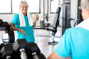 Smiling and active retired senior woman in gym area takes a break from exercises drinking. Sport,...
