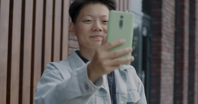 Carefree Asian guy taking selfie with thumbs-up hand gesture posing for smartphone camera outside in city street. Gadgets and photography concept.