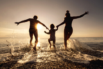 Family of mom, dad and young daughter runs to sunset beach with lots of splashes