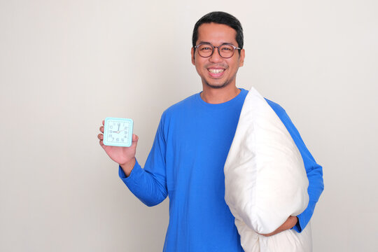 A man holding sleeping pillow and showing alarm clock with happy expression