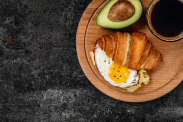 Croissant sandwich with avocado, cheese and egg and coffee cup on black background.