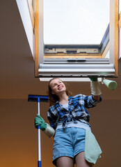 Cheerful young woman cleans skylight window in her apartment. Housework, cleaning window at home.