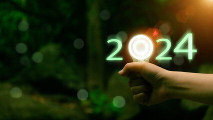 New Year concept. Hand holding light bulb with 2024 icon with goals, nature, morning sunlight....