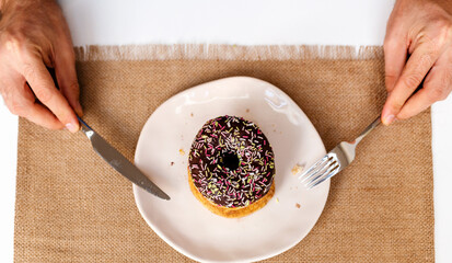 High angle view of chocolate donut with sprinkles on plate. Doughnut on plate and fork and knife in hands. - Powered by Adobe