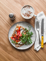 Delicious breakfast, brunch -  crepes with lightly salted salmon, cream cheese, arugula and cappuccino on a wooden background, top view