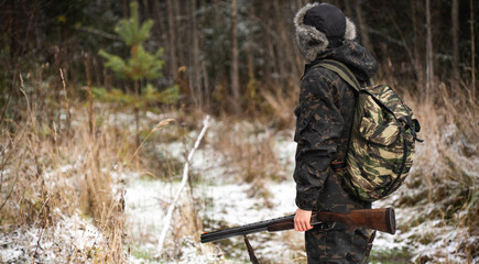 Male hunter in camouflage and with backpack, armed with a rifle, walks through the snowy winter...