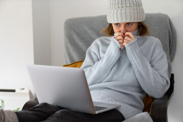 A woman in warm clothes with laptop. Low heating temperature in the house. Energy crisis concept.