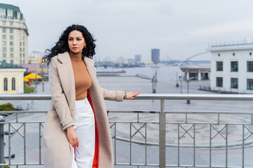 A beautiful woman with black curly hair poses leaning on a bridge with the city and the river in the background. The woman is sad and looking away. The concept of the city, rest and work, thoughts