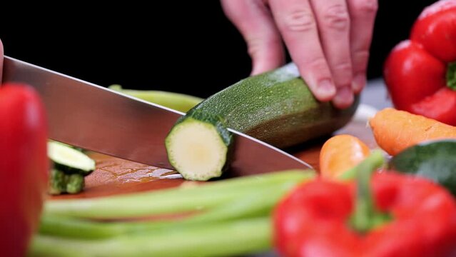 Person with sharp knife cuts zuccini into slices between various fresh vegetables peppers, carrots, leeks on cutting board