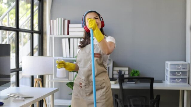 Happy young woman listening to music and dancing while mopping the floor, cleaning the home office.