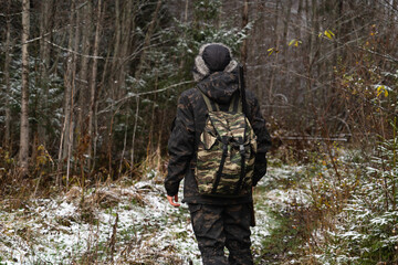 Male hunter in camouflage and with backpack, armed with a rifle, walks through the snowy winter...