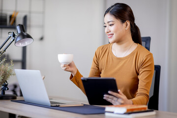 A beautiful young asian woman holding cup and working on laptop computer while drinking coffee at home. concept of freelancer planning Business finance documents financial.