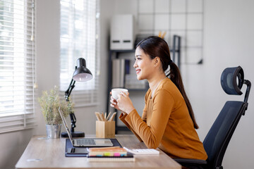 A beautiful young asian woman holding cup and working on laptop computer while drinking coffee at...