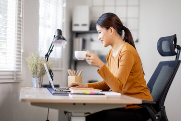 A beautiful young asian woman holding cup and working on laptop computer while drinking coffee at...