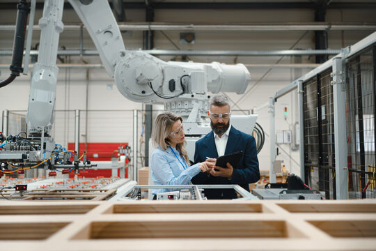 Female engineer and male project manager standing in modern industrial factory, talking about production of wooden furniture. Big furniture manufacturing facility with robotics and automation.