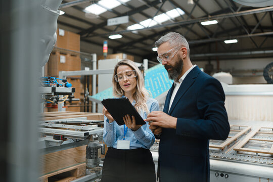 Female engineer and male project manager standing in modern industrial factory, talking about production of wooden furniture. Big furniture manufacturing facility with robotics and automation.