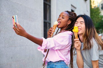 Two multiracial happy friends with ice creams taking a selfie in summer at street.