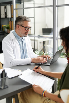 Vertical image of mature doctor in white coat typing on laptop while having consultation with woman in office
