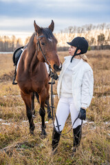 Beautiful blond professional female jockey standing near horse in field in winter. Friendship with horse concept