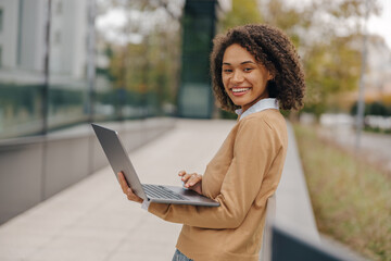Pretty female entrepreneur working on laptop standing outside on modern building background
