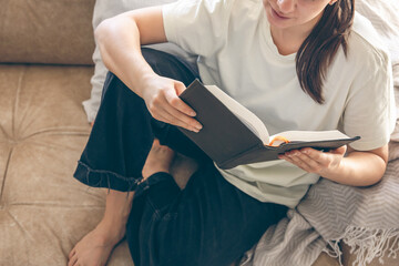 Young Caucasian woman reading a book, sitting on the sofa at home.