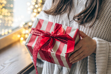 A holiday gift with a red bow in female hands on a blurred one with garlands.