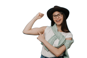 Adorable young brunette girl dressed in white t-shirt, brown hat and knitted sweater tied around neck, shows her biceps and points at it by another hand with wide smile against transparent background
