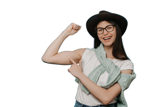 Adorable young brunette girl dressed in white t-shirt, brown hat and knitted sweater tied around neck, shows her biceps and points at it by another hand with wide smile against transparent background