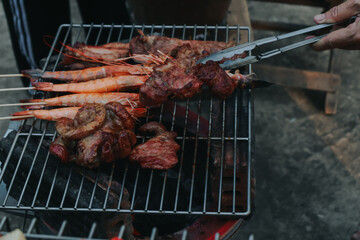 People are cooking seafood and vegetable skewers on a charcoal grill. with a plate of shrimp placed next to it A warm family holiday celebration. BBQ party