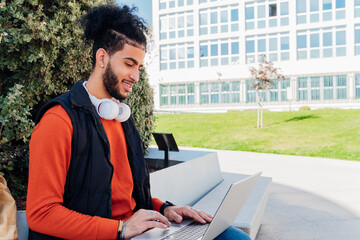 Side view of a student with casual clothes and heapdhones using laptop in the campus
