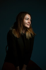 Classic dark studio portrait of a young brown-haired woman in black clothes sitting on a chair.